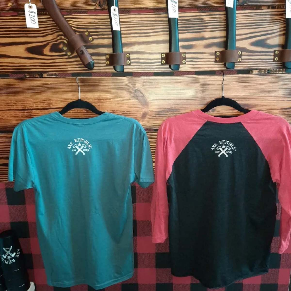 Two shirts hanging under mounted axes on a wooden wall.
