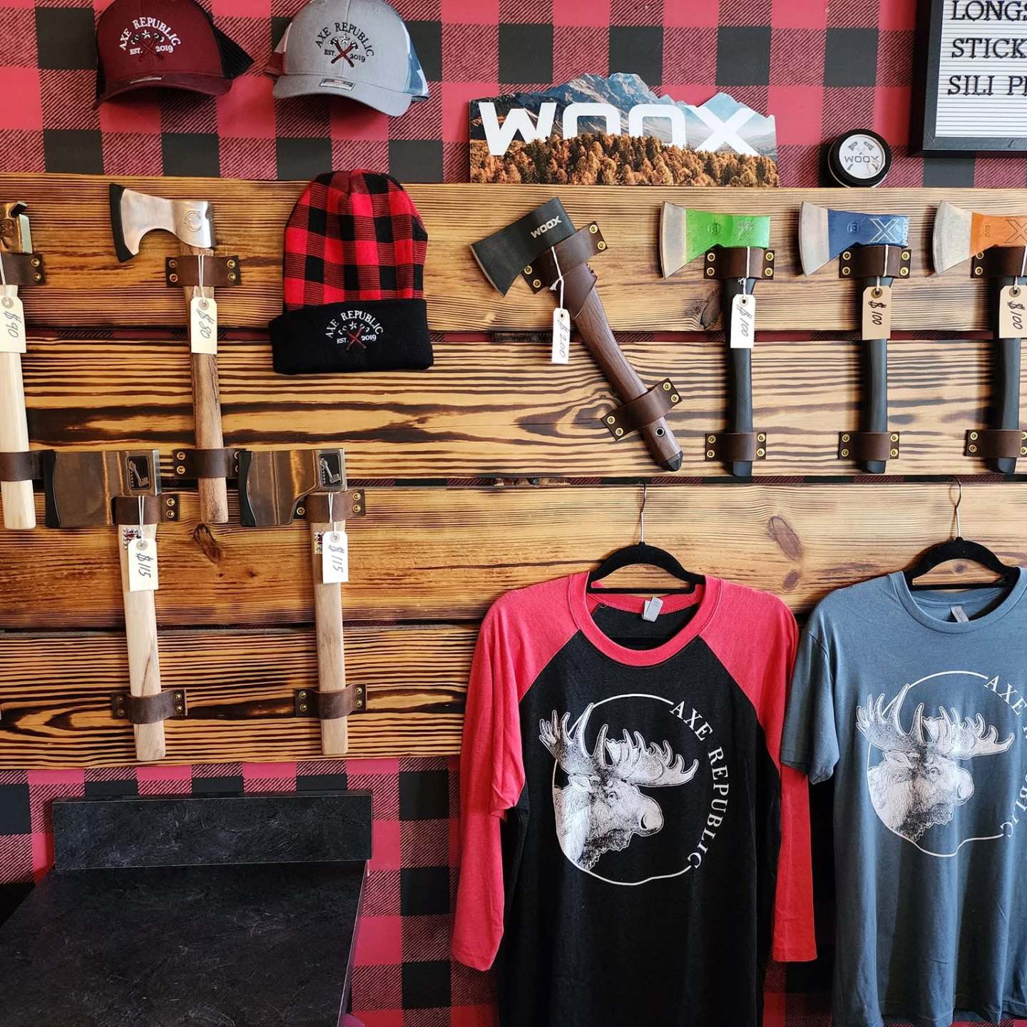 Display of axes, hats, and shirts with moose design on a plaid wall.