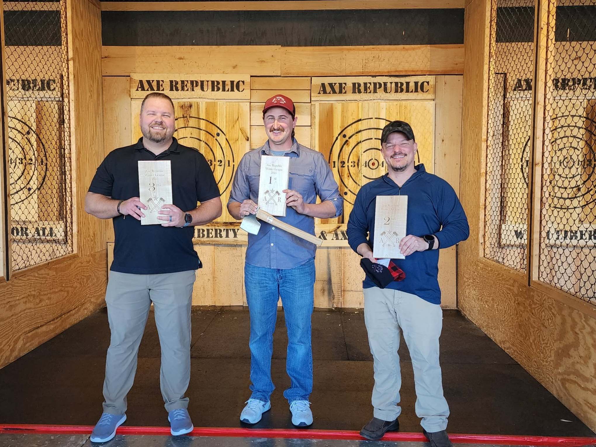Three men holding awards in front of an axe-throwing target backdrop.