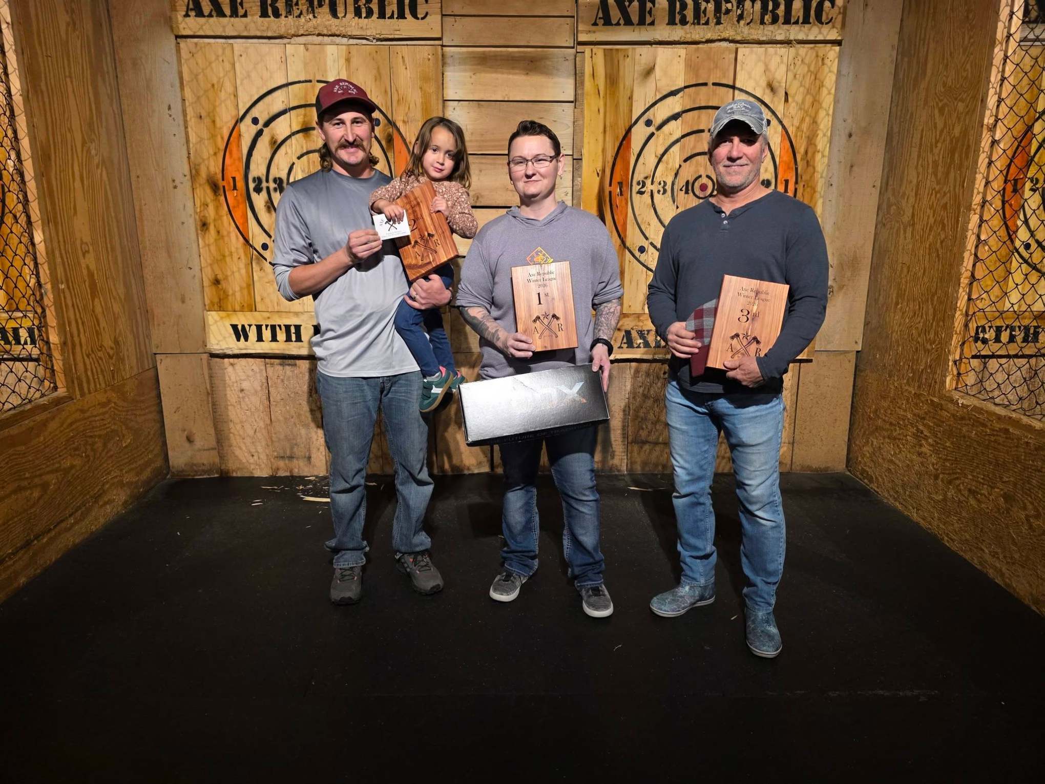 Three people holding awards in front of axe throwing targets.