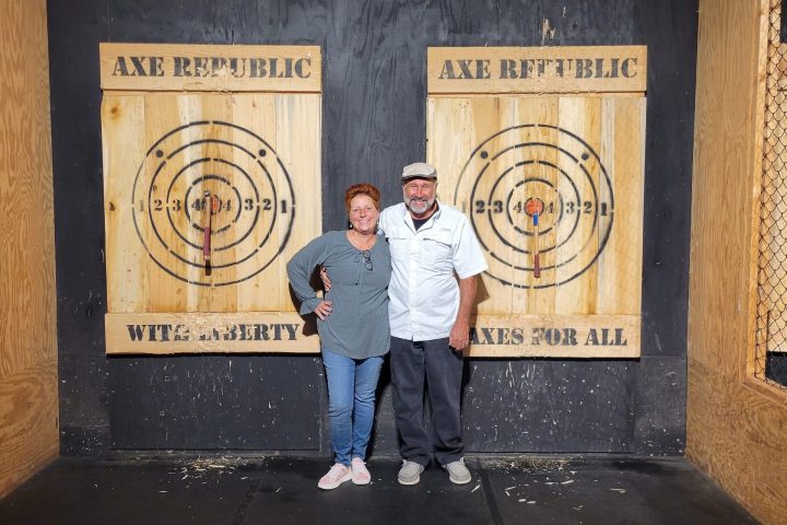 Two people posing in front of wooden axe throwing targets indoors.