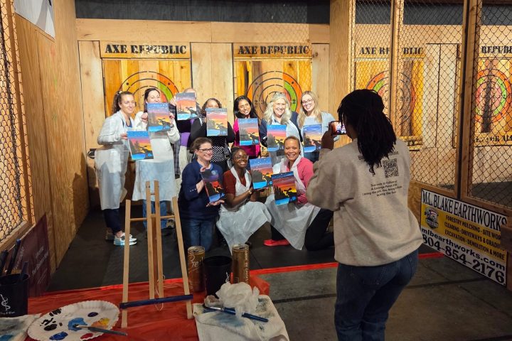 Group of people posing with paintings in an indoor venue with painting supplies and wooden backdrop.