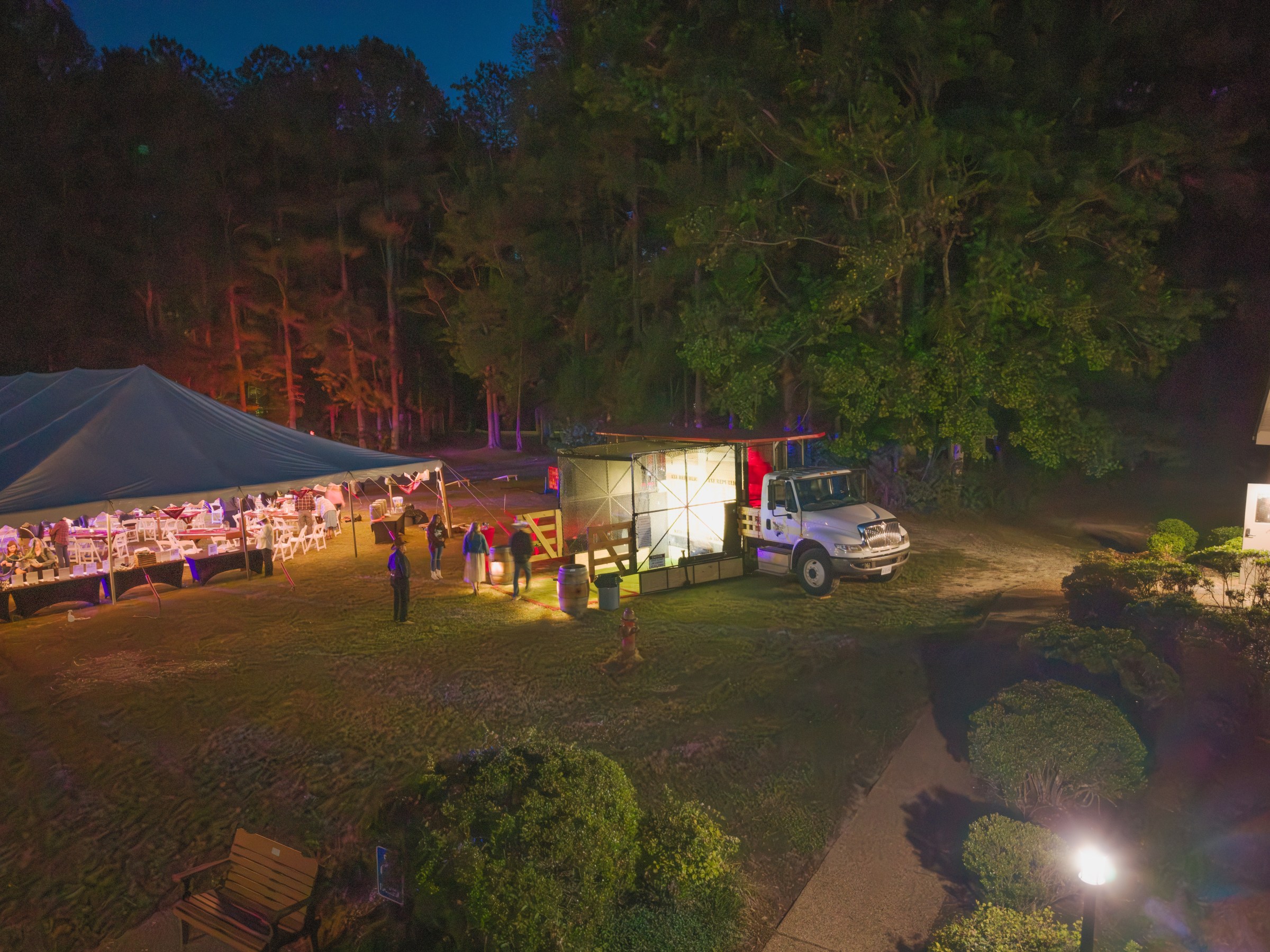 Outdoor evening event with a tent, truck, and illuminated area under trees.